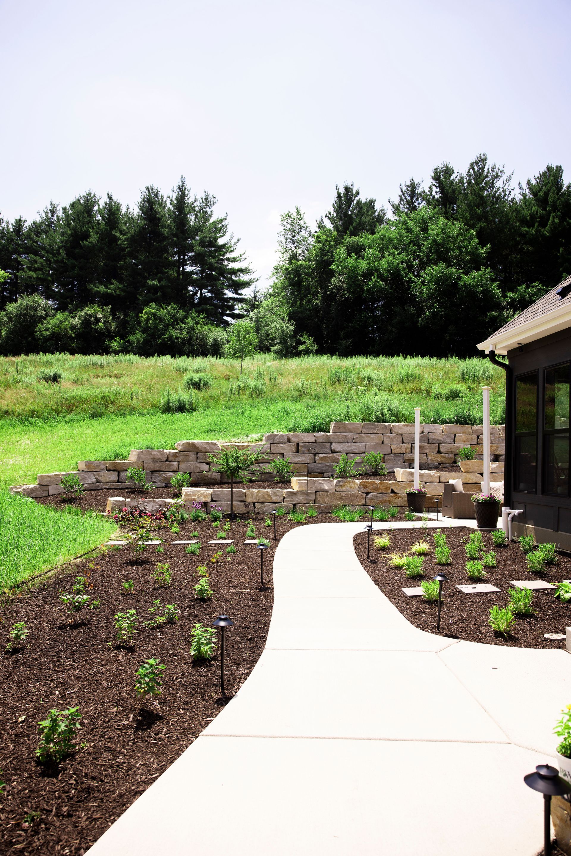 Pathway curving through a landscaped yard with a stone retaining wall and lush greenery, leading toward trees under a bright sky.