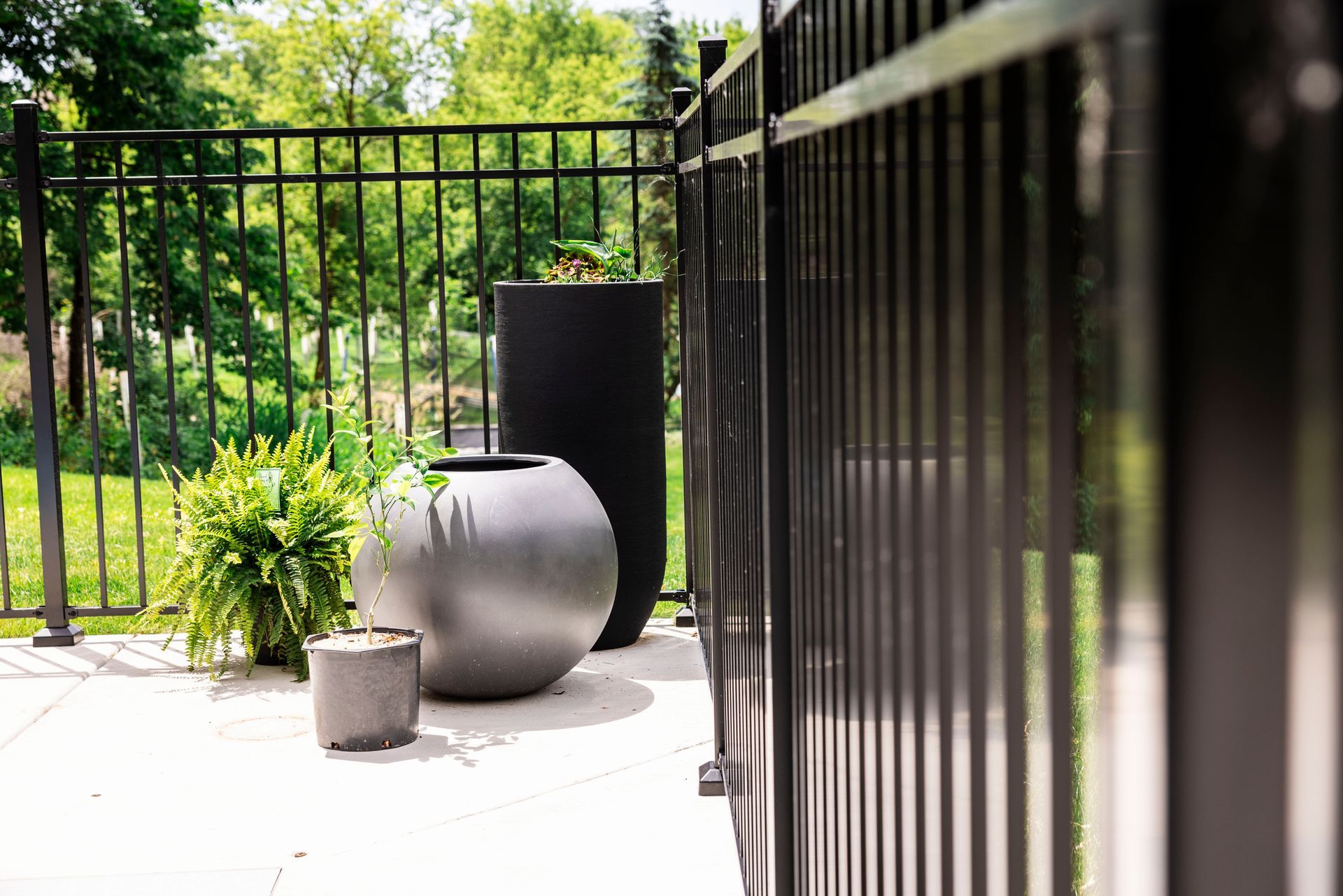 Black iron fence bordering a patio with gray planters and greenery, trees in the background.