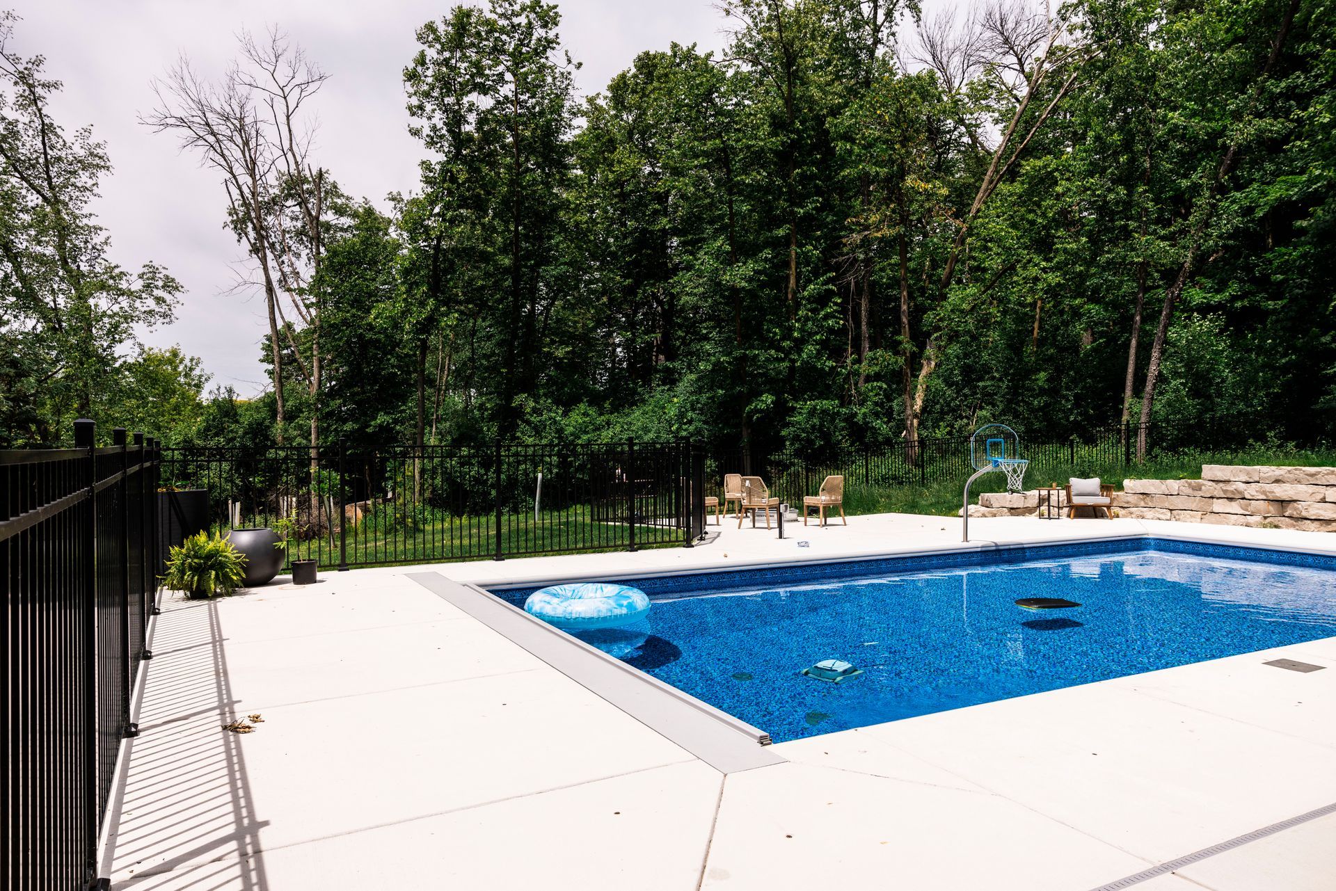 Swimming pool with blue water and white patio, surrounded by black fence and trees.