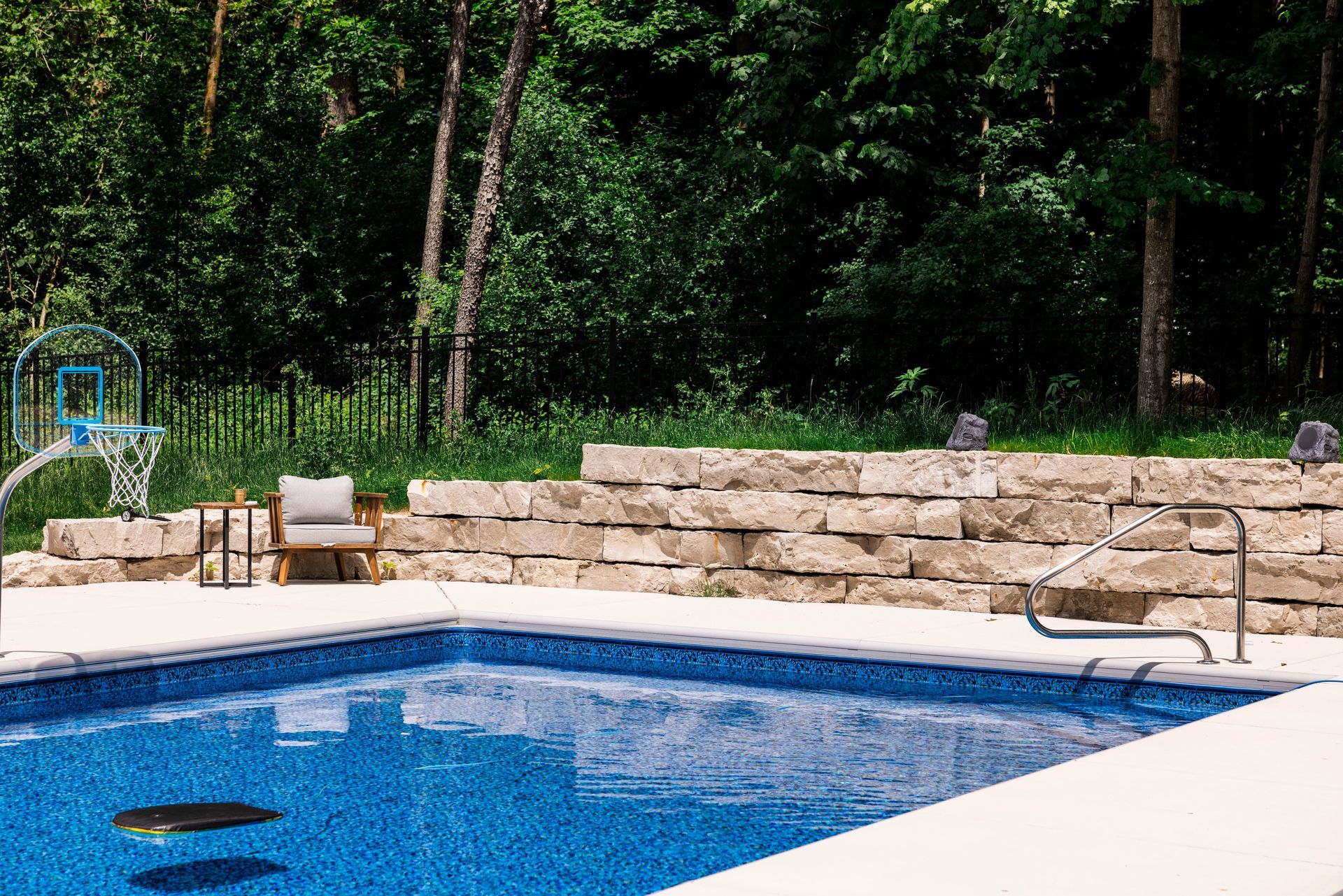 Swimming pool with blue water and stone wall, basketball hoop, and trees.