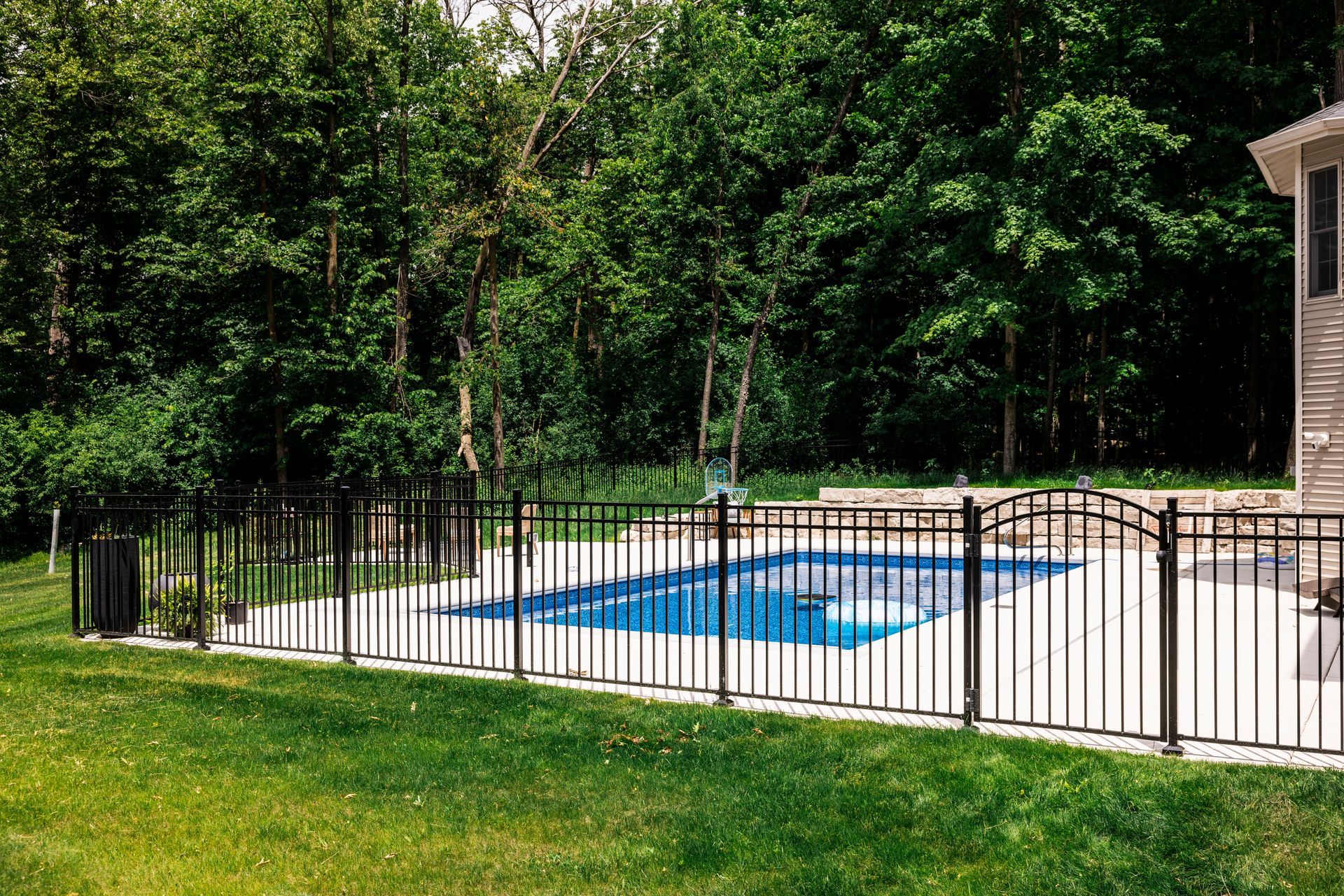 Black fenced-in rectangular pool with blue water on a concrete patio in a backyard with green grass and trees.