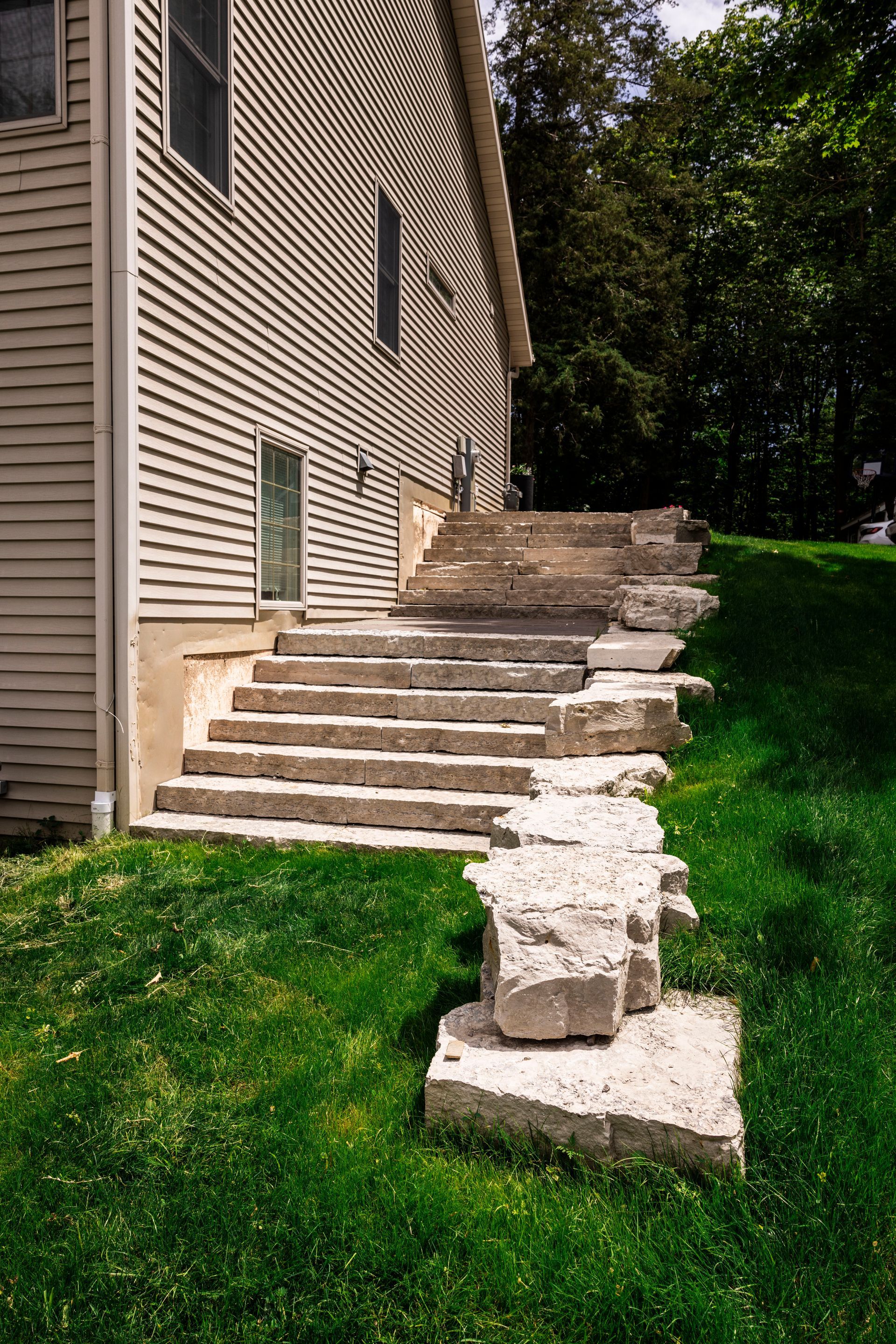 Concrete steps lead up a grassy hillside next to a house with beige siding.