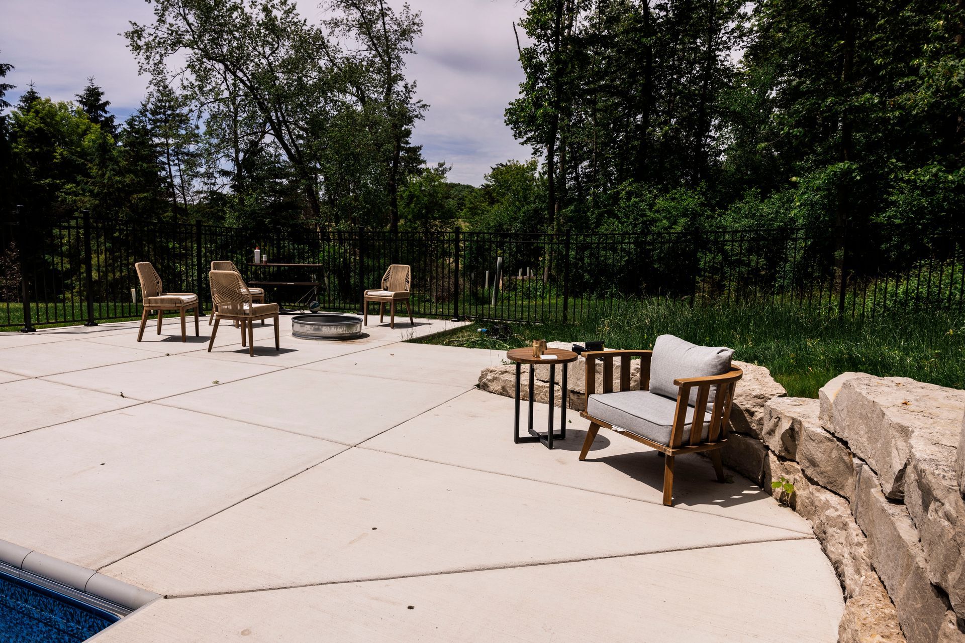 Outdoor patio with seating, fire pit, and lush greenery in the background. Sunny day.