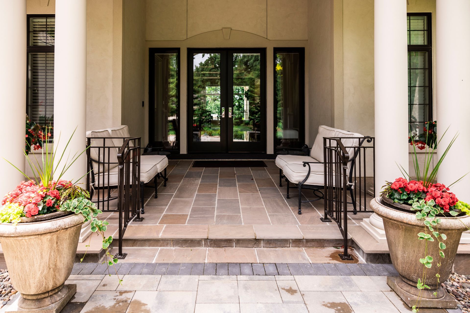 Entrance with double doors, stone patio, columns, two benches, flower pots.