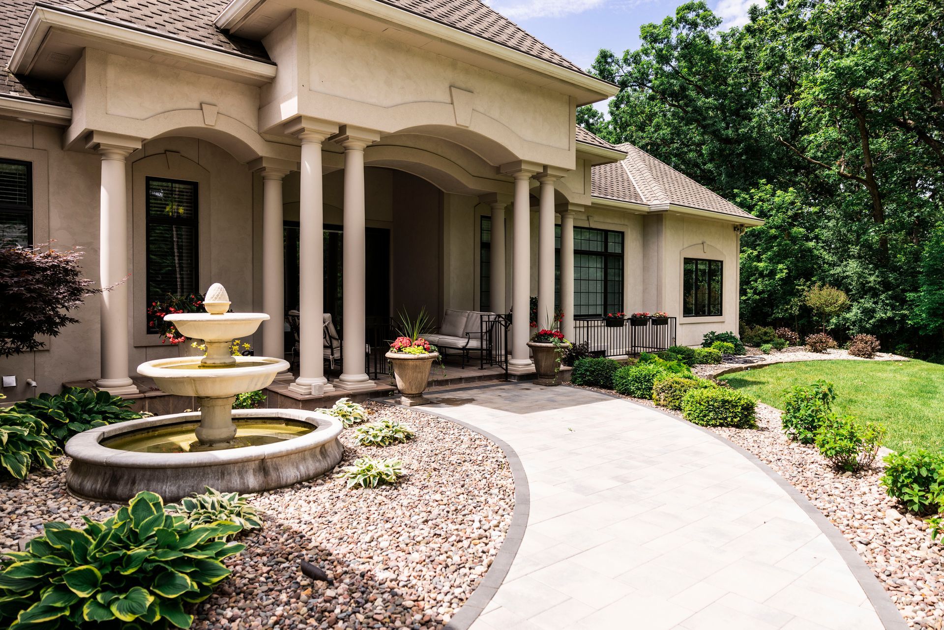 Elegant home with colonnade entrance, fountain, and paved walkway leading to the front door. Lush landscaping.
