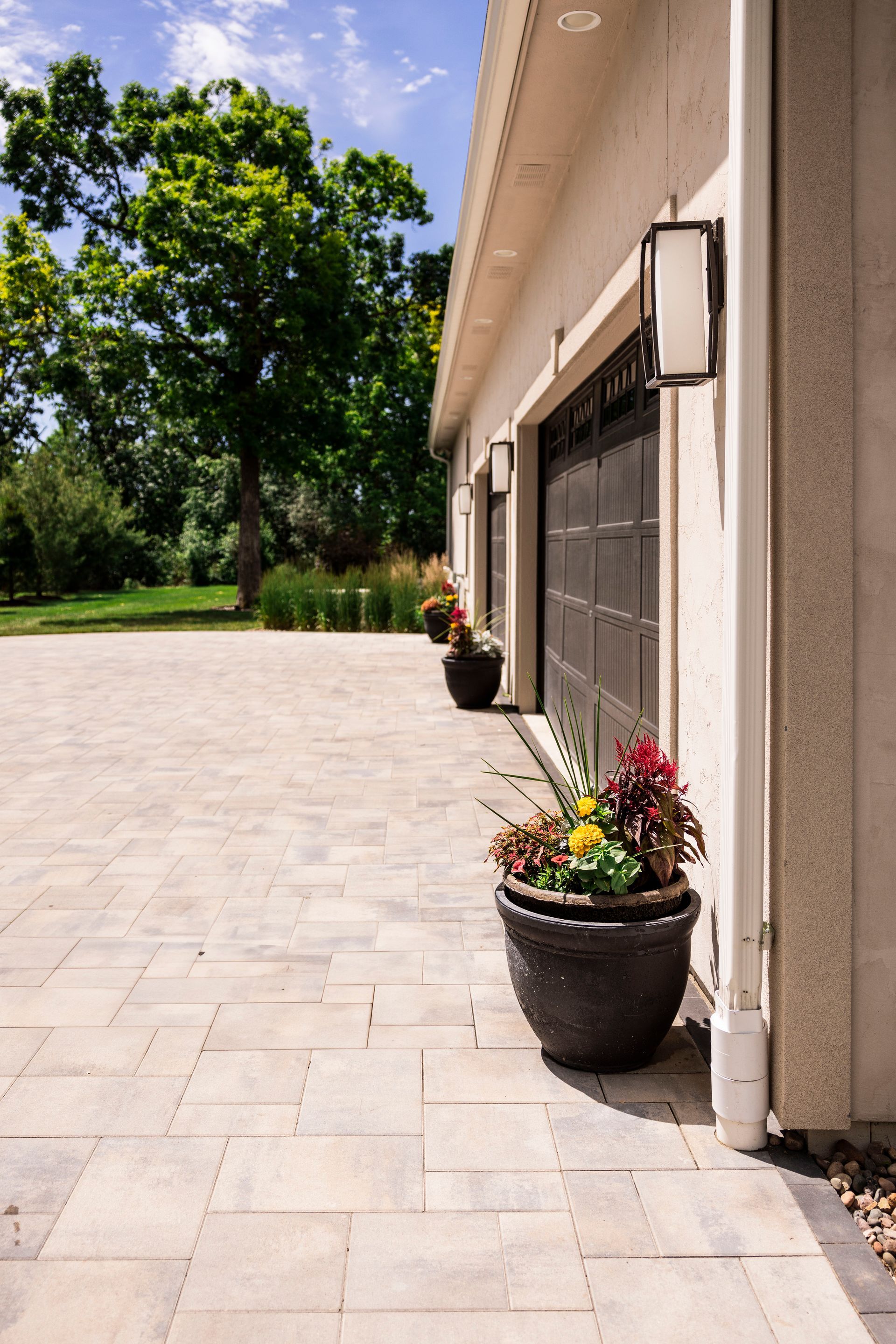 Garage exterior with brick pavers, flower pots, and trees on a sunny day.