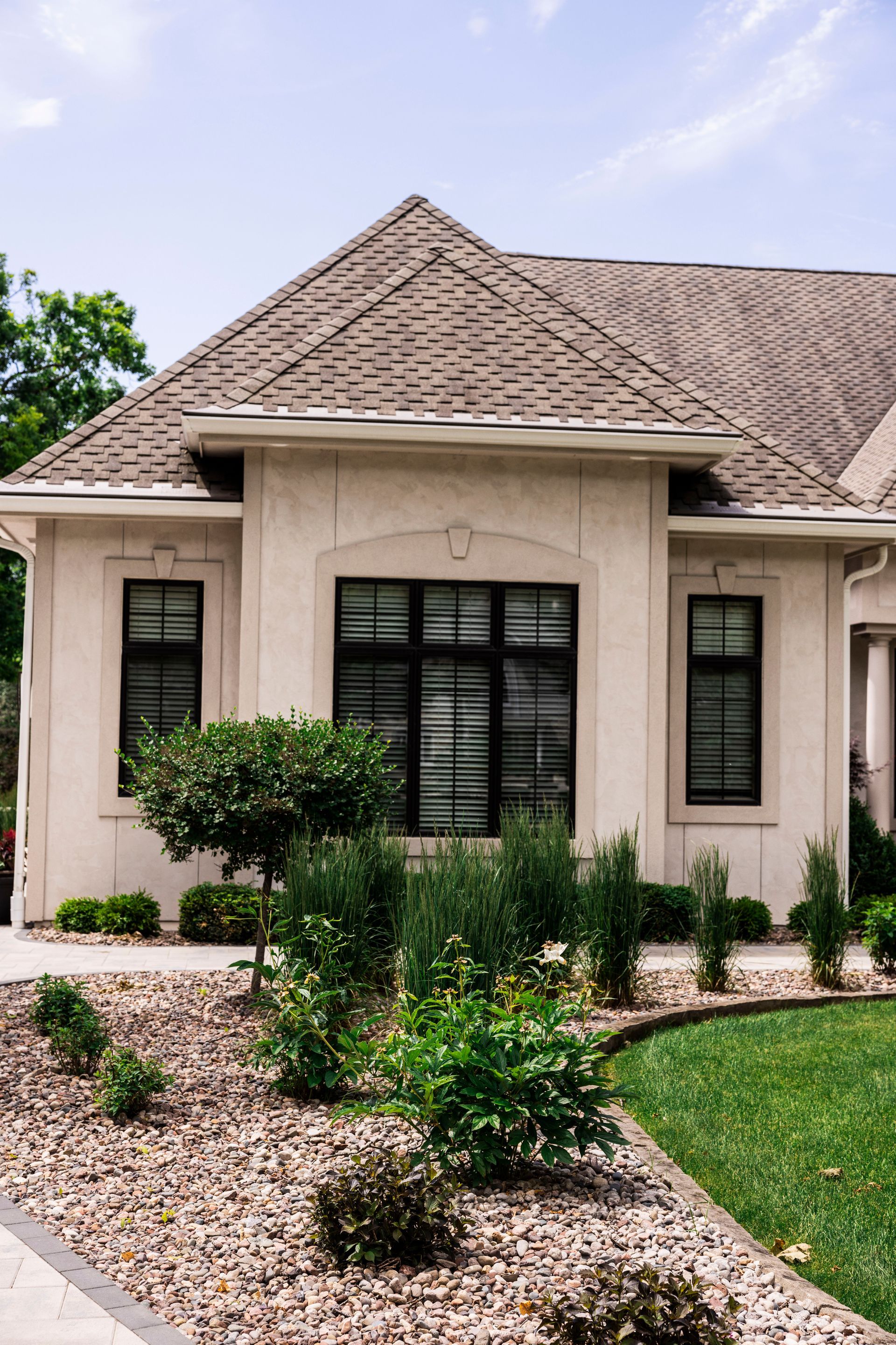 Beige house with a brown shingle roof and black framed windows; landscaped yard with green grass and shrubbery.