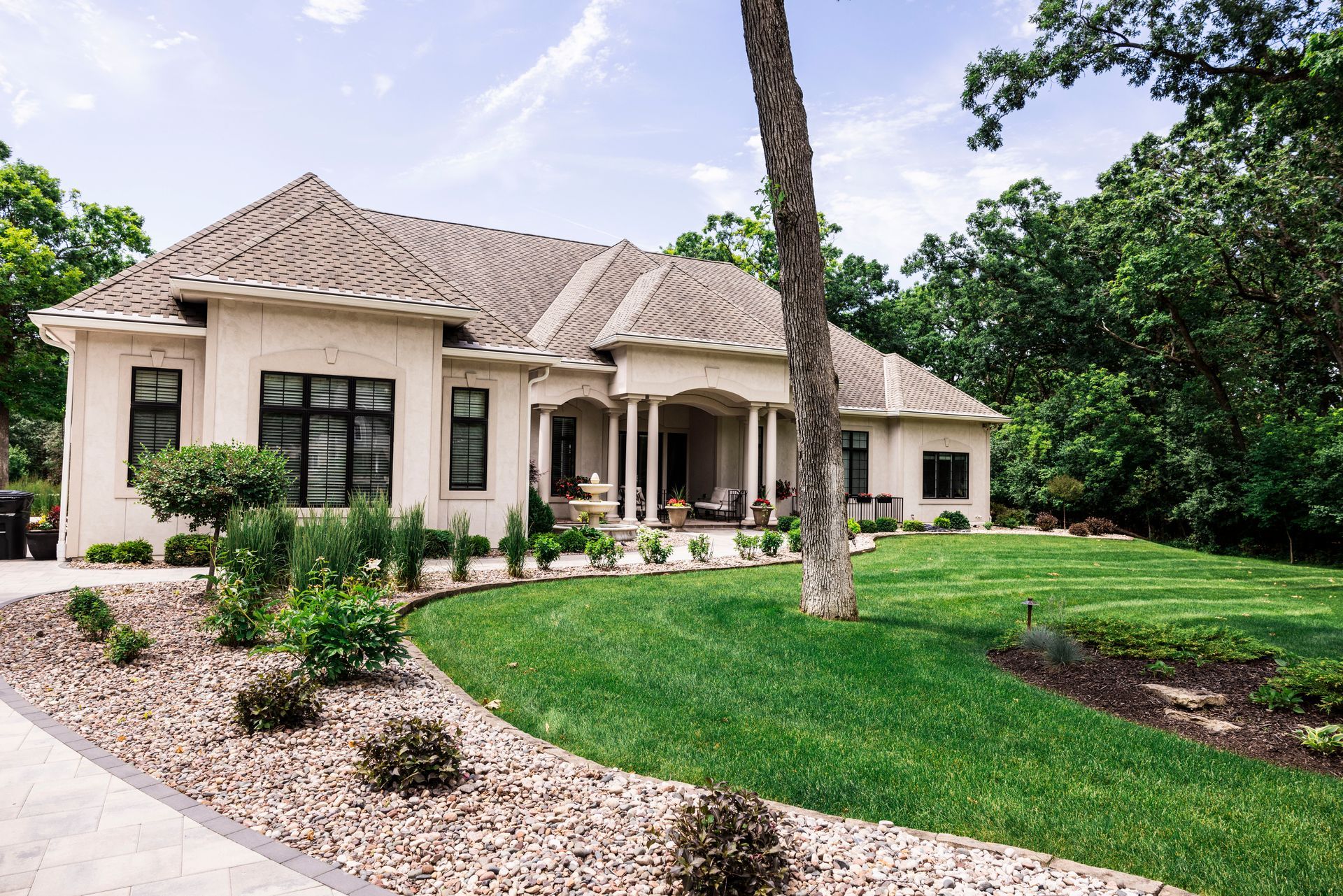 Beige house with manicured lawn, stone pathway, and trees under a blue sky.