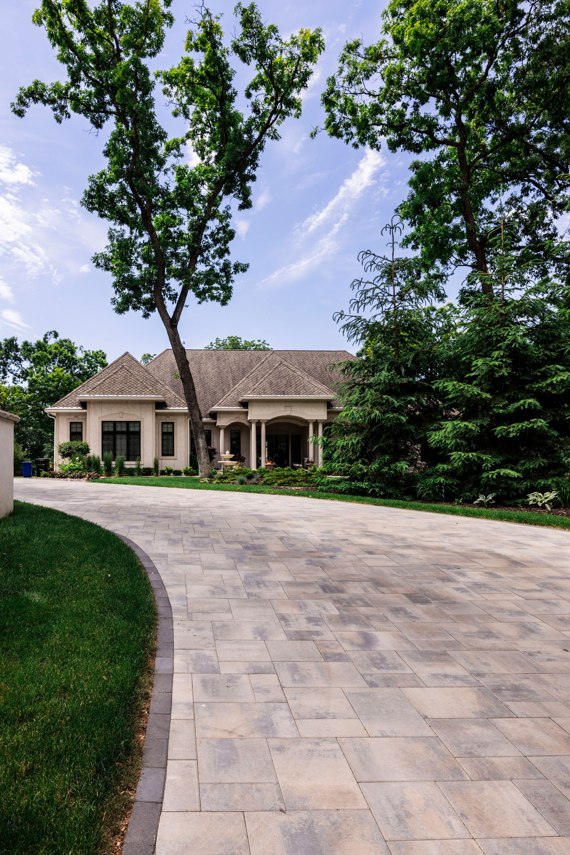 Paved driveway curves toward a beige house with trees on a sunny day.