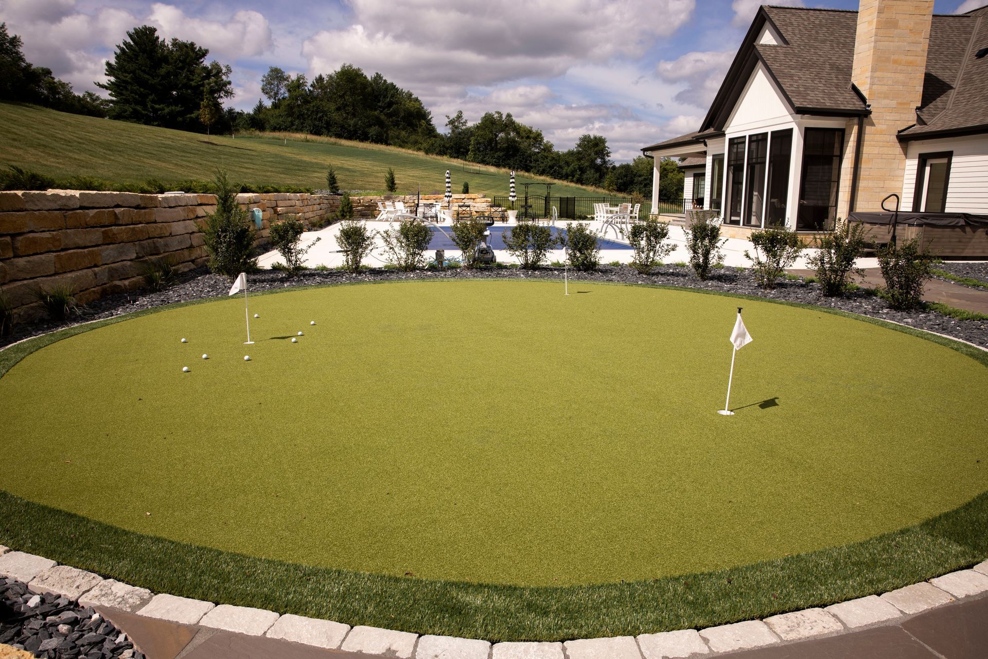Circular green artificial putting green, flagsticks, bordering stone, house in background.