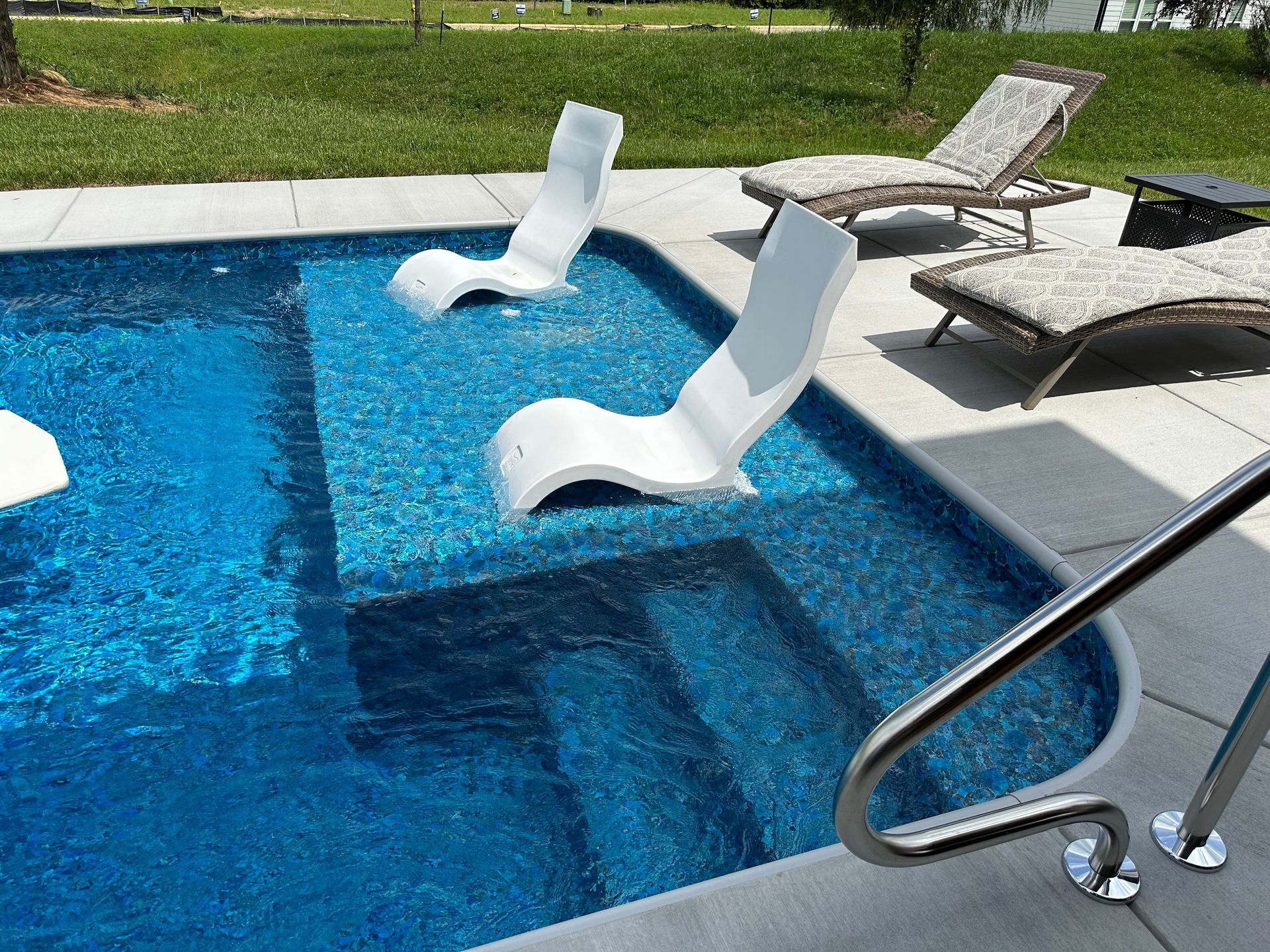 Pool with two submerged white lounge chairs, stairs, and deck chairs. Sparkling blue water, sunny outdoor setting.