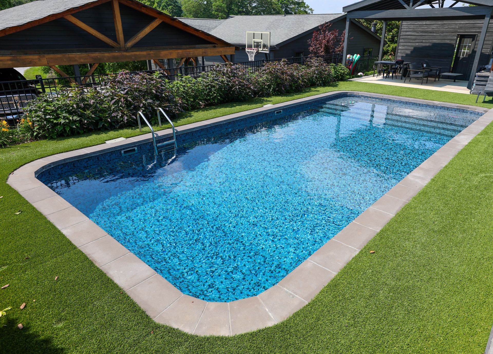 Rectangular swimming pool with blue mosaic tile, surrounded by green artificial grass and a stone border.