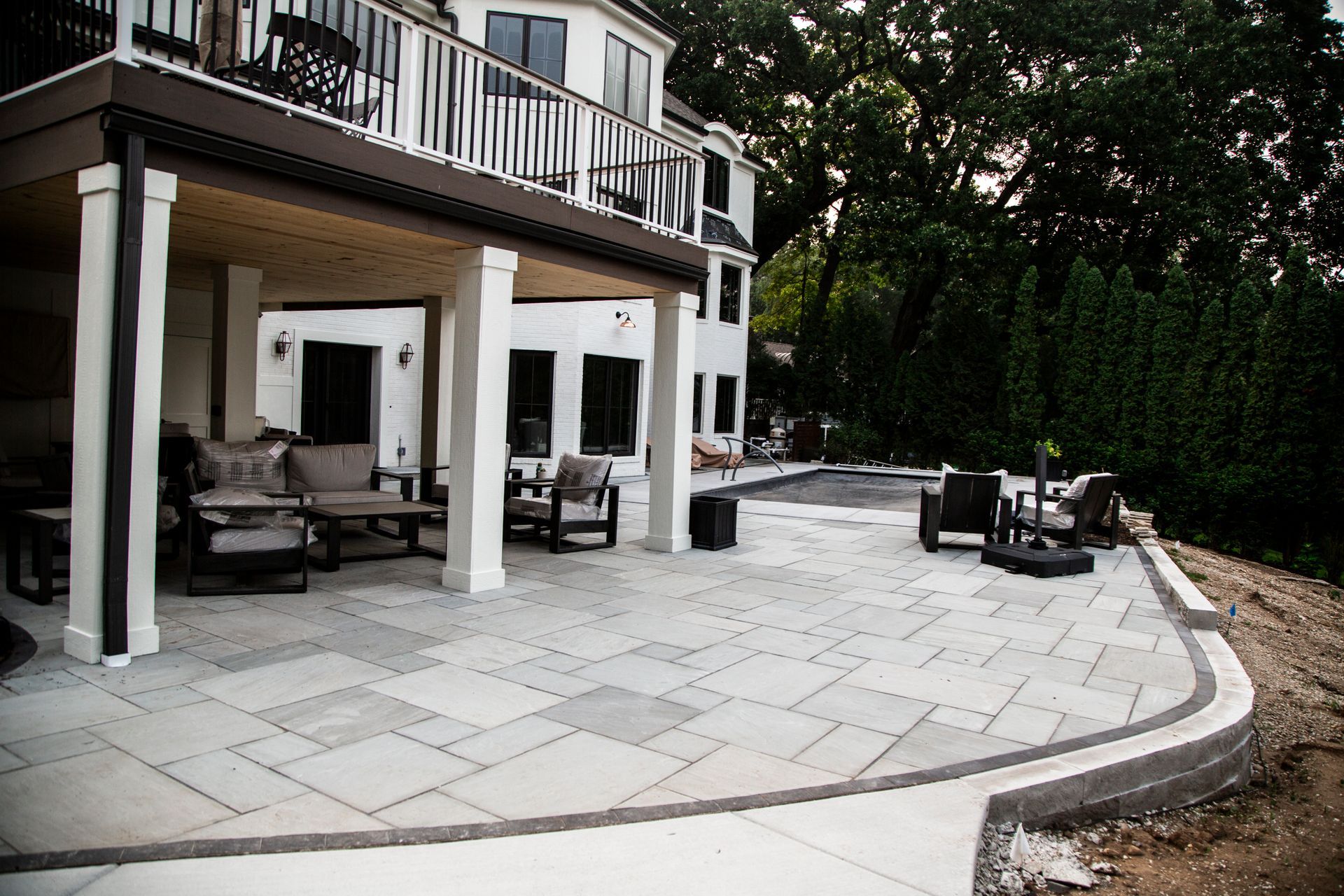 Stone patio with outdoor furniture under a white house balcony near a pool.