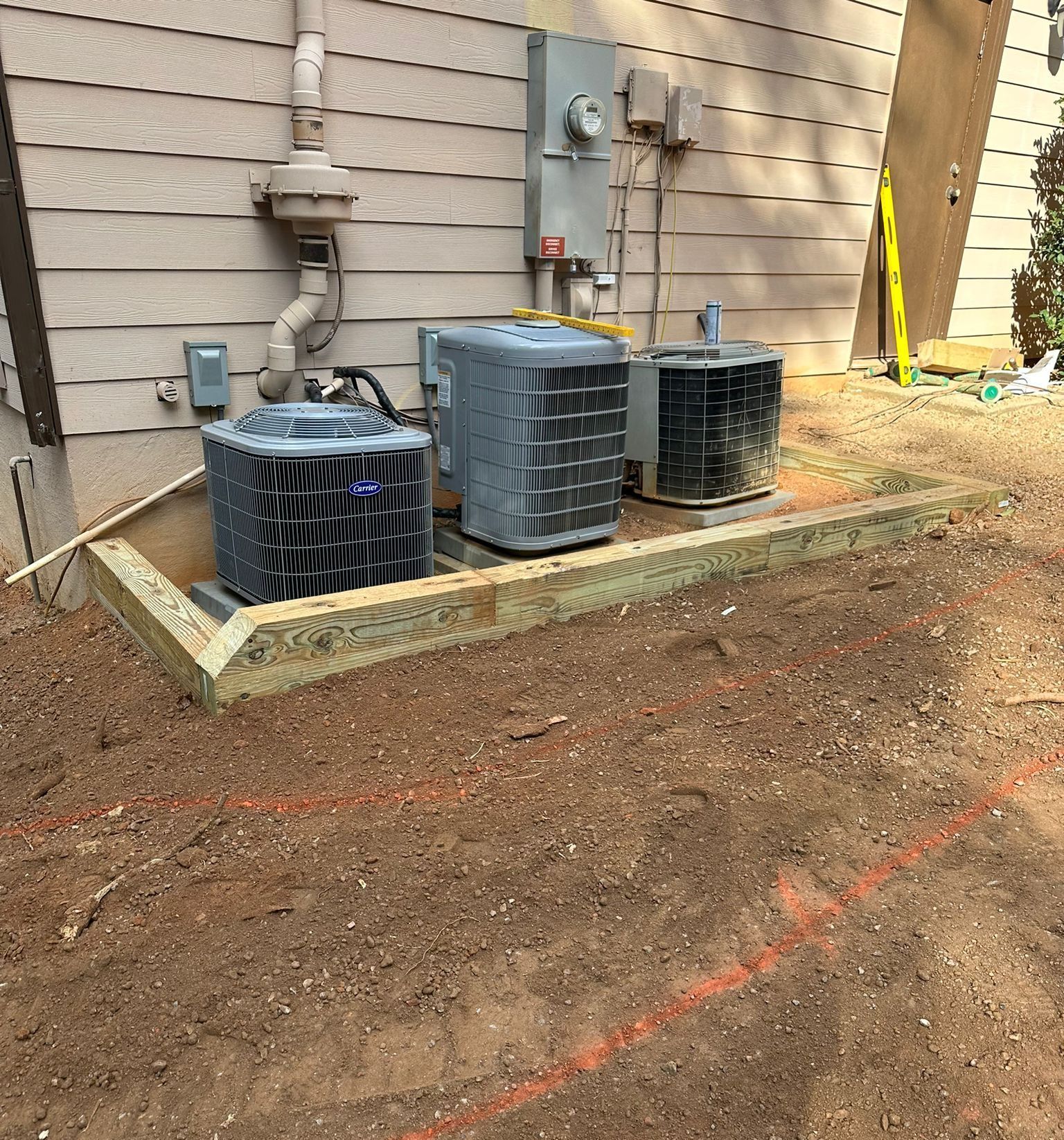 Two air conditioners are sitting in the dirt in front of a house.