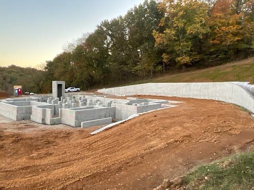 Construction site with concrete foundation and retaining wall, brown earth, and trees.