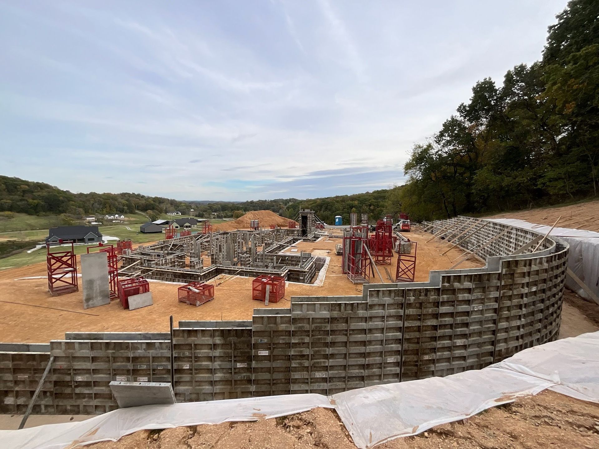 Construction site with curved retaining wall and concrete foundation. Red scaffolding and earthworks visible under a cloudy sky.