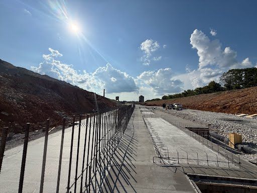 Construction site with concrete road, rebar, and earth embankments under a bright blue sky.