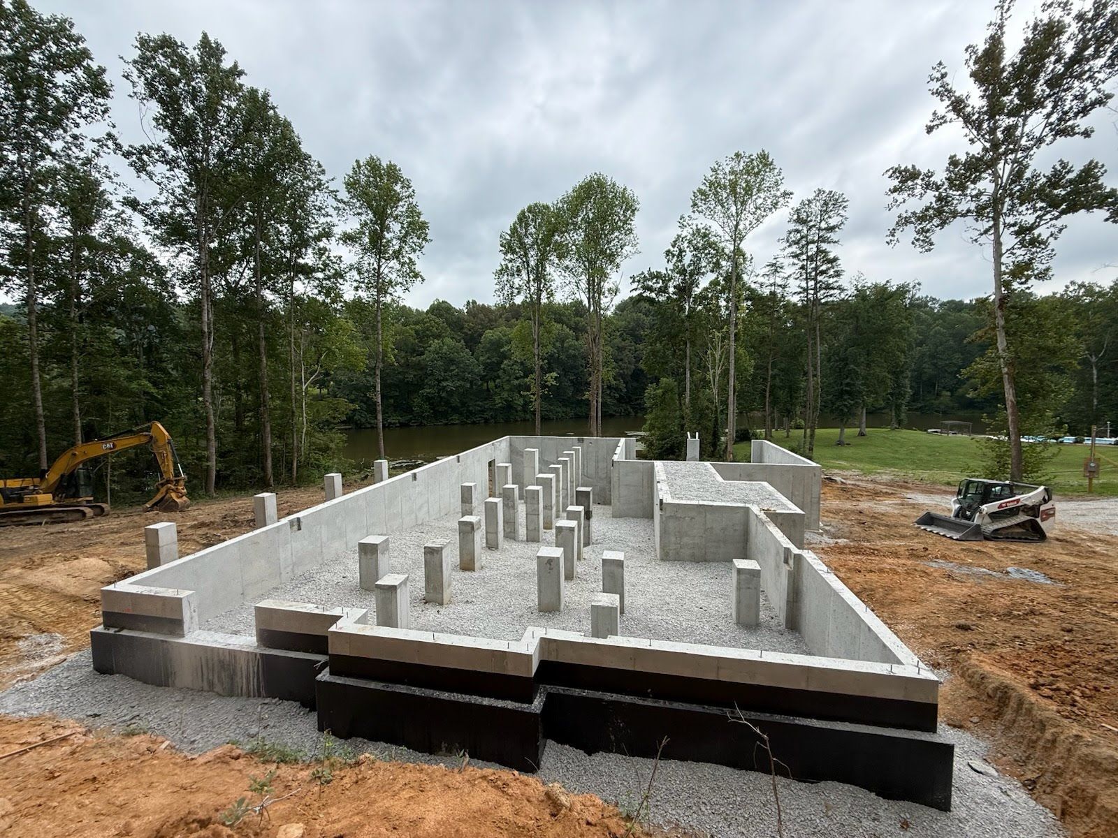 Foundation of a building under construction; concrete walls and support pillars in a dirt setting, trees in the background.