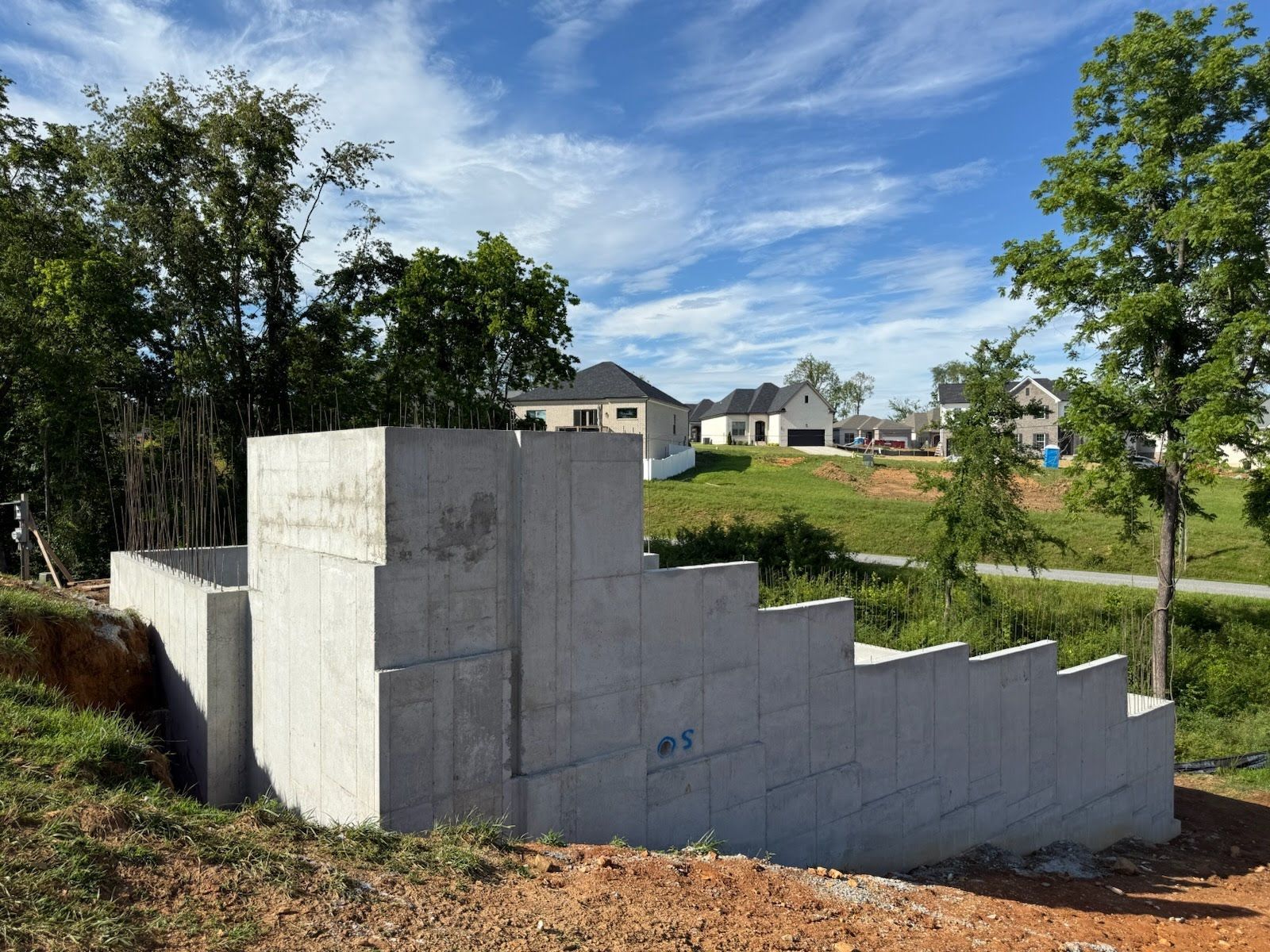 Concrete retaining wall under construction with stepped design, in a grassy area with houses in the background.