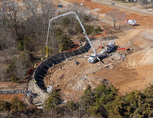 An aerial view shows a concrete pump truck pouring concrete into a curved retaining wall at a construction site.