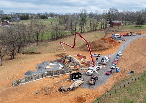 A construction site in a field with a red concrete pump truck pouring concrete into a foundation near parked vehicles.