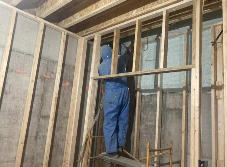 A man in blue overalls is standing on a scaffolding in front of a wooden wall.