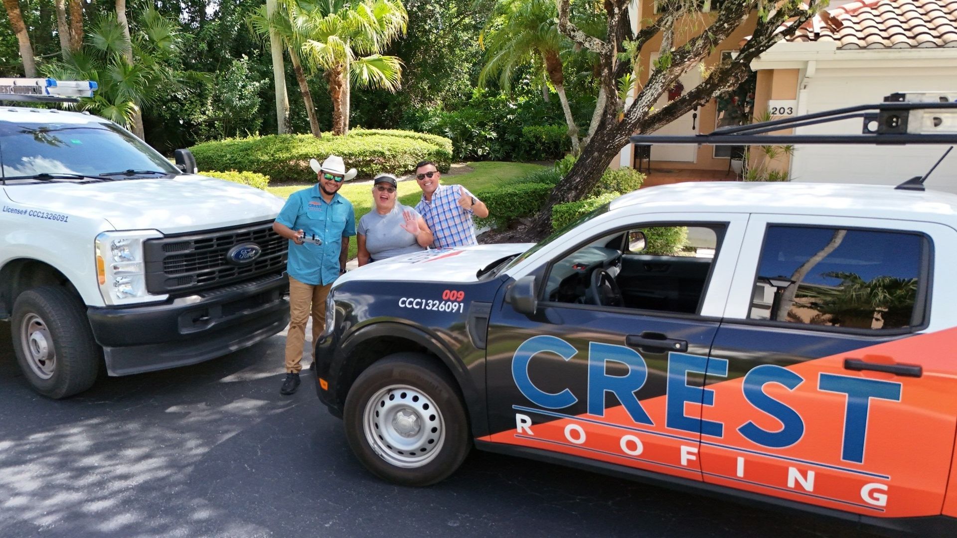 Three people smiling next to Crest Roofing trucks in a driveway. Trees and house in the background.