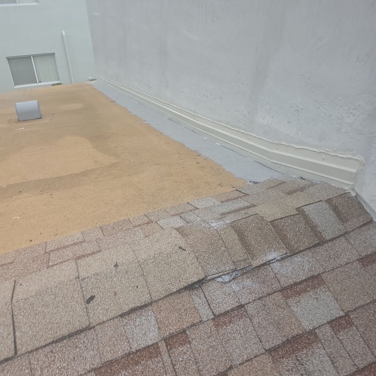 Close-up of a roof where shingles meet a wall; the roof is brown, the wall is white, and the area between has gray sealant.