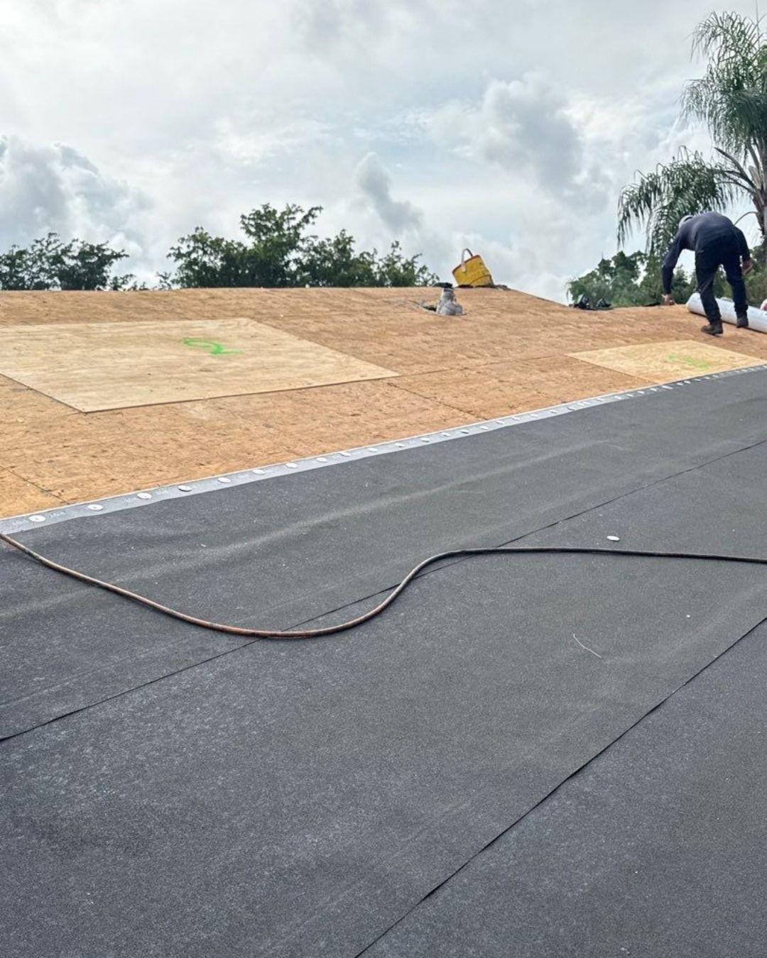 Roofer installing a black flat roof. Brown wood substrate visible. Cloudy sky.