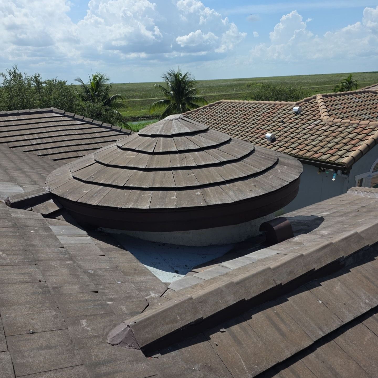 Dome-shaped skylight on a tile roof, surrounded by other roofs and greenery against a blue sky.