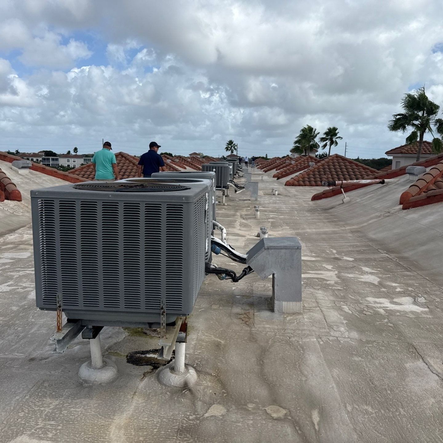 Two people on a flat roof with several air conditioning units. Palm trees and other buildings in the distance, cloudy sky.