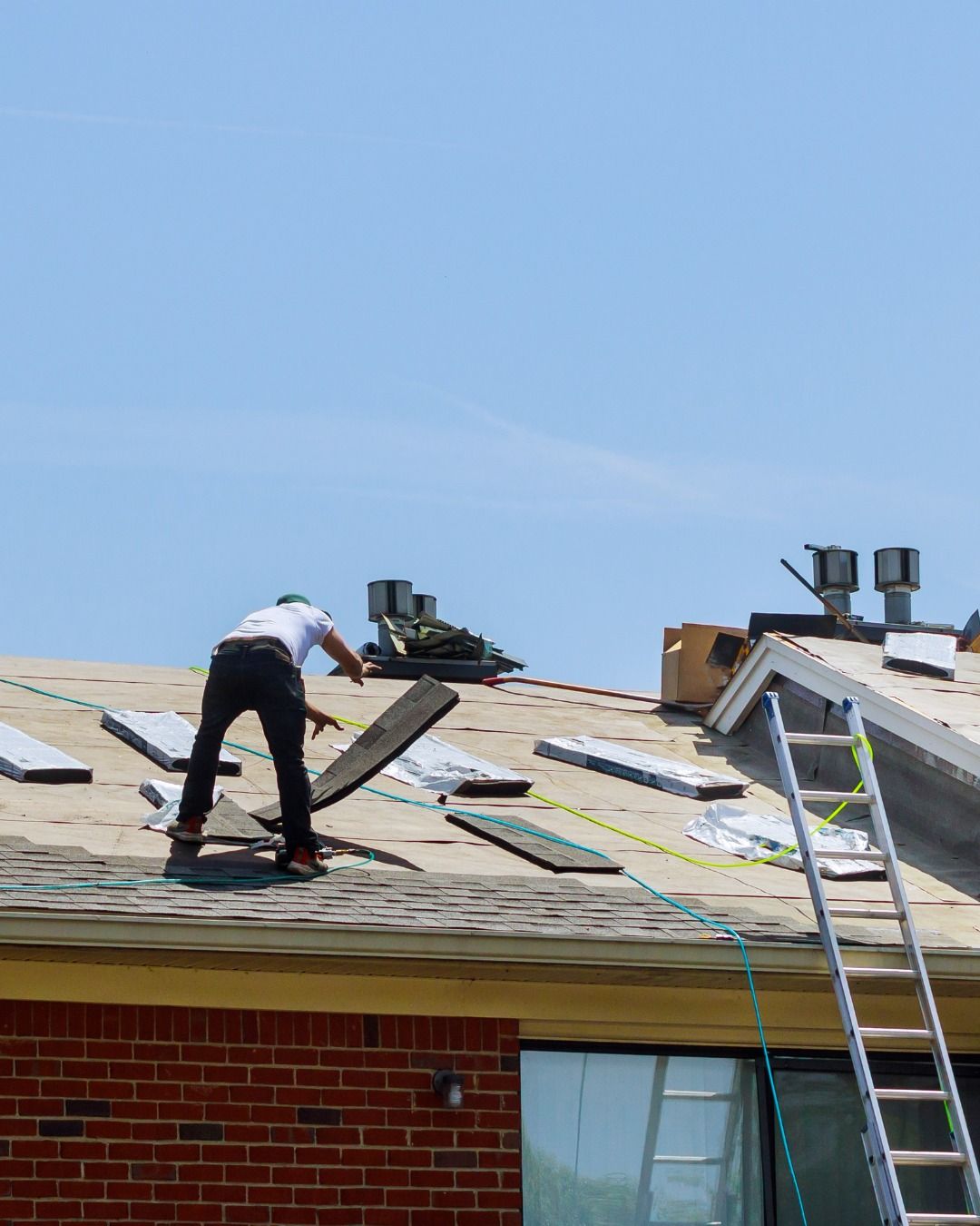 Roofer on a sunny day, replacing shingles. Ladder leans against roof.