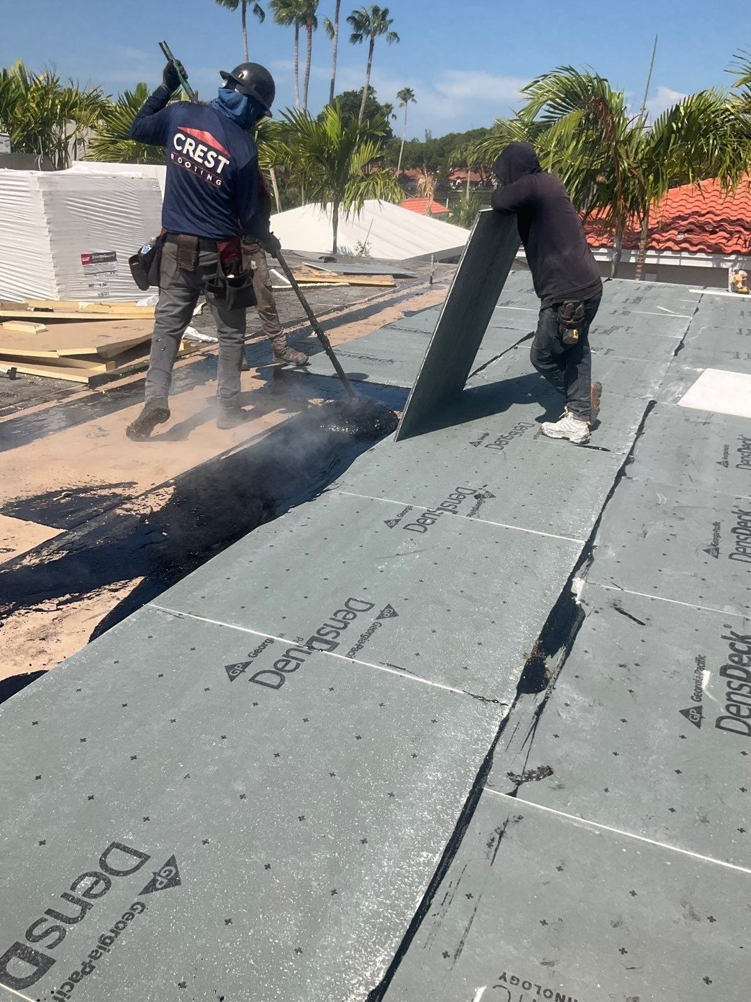 Two roofers installing roofing material on a sunny day. One uses a torch, the other positions a panel.