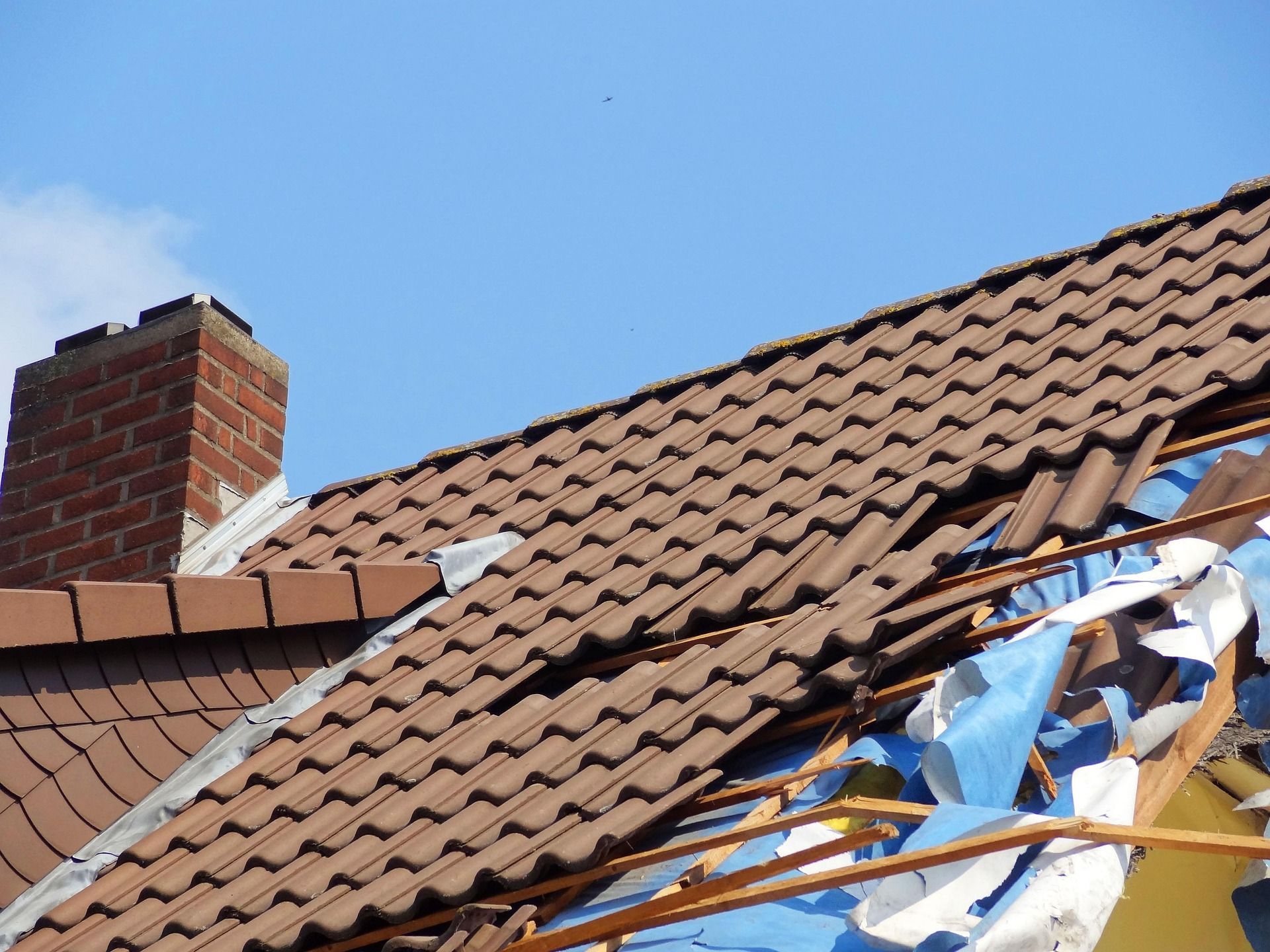 Damaged brown tiled roof with exposed rafters and a brick chimney against a blue sky.