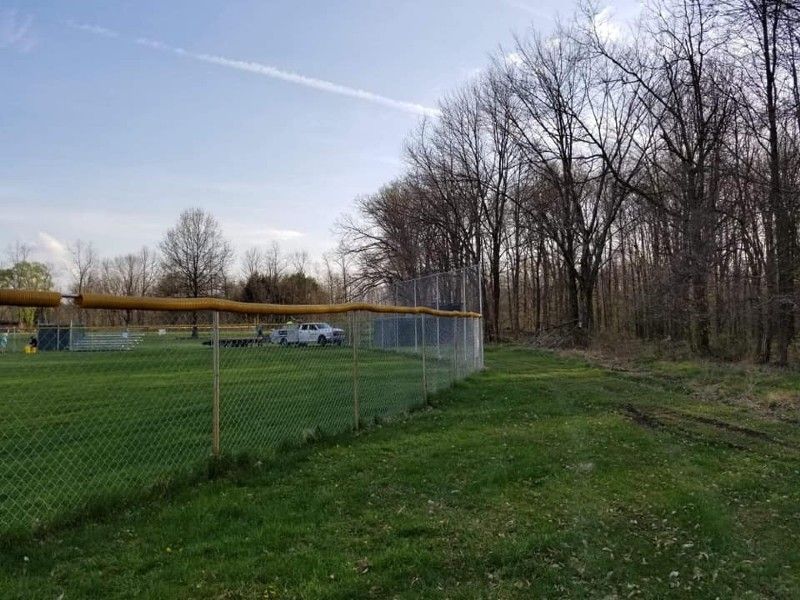 Baseball field with a chain-link fence, green grass, and trees. A vehicle is parked on the field.