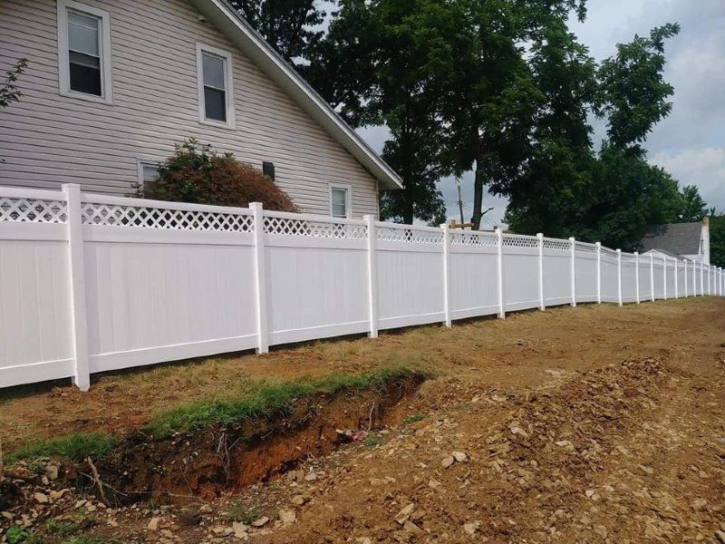 White vinyl fence surrounding a house; a trench is dug along the fence line in the dirt.
