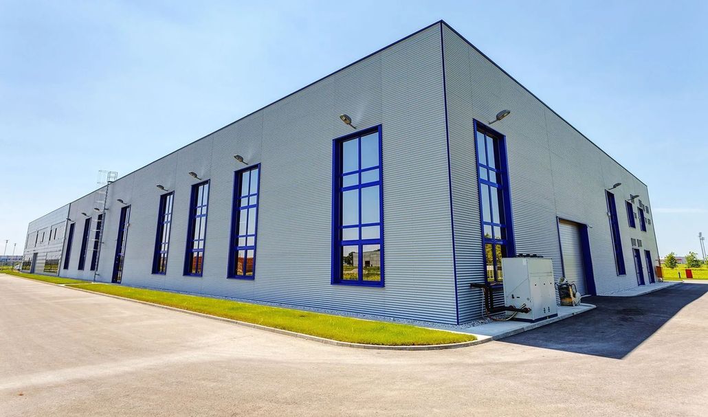 Large gray industrial warehouse with blue-trimmed windows on a sunny street corner