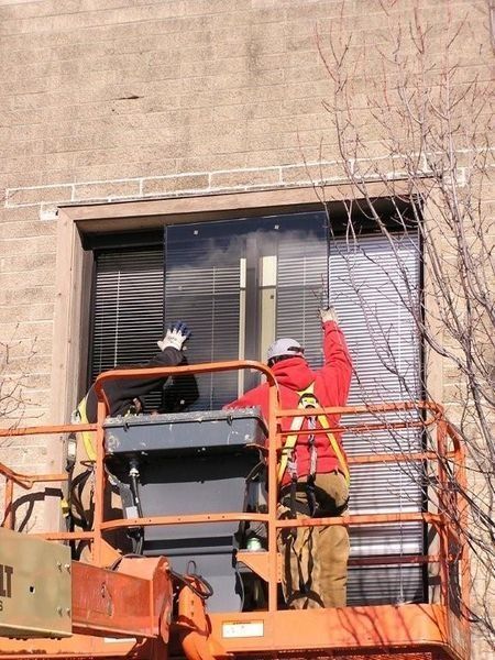 Two workers in orange lift basket on an exterior building, one waving beside a window.