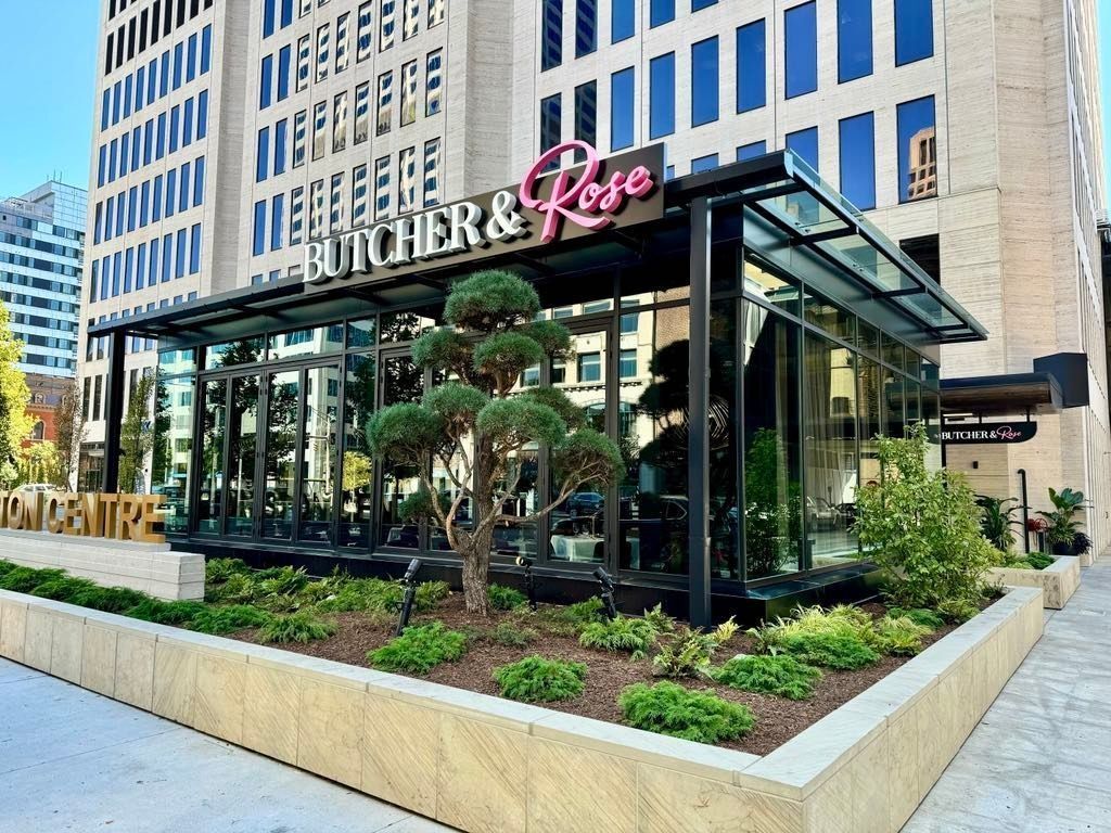 Exterior of a modern glass storefront with a pink-and-white sign, landscaped planter, and office buildings behind
