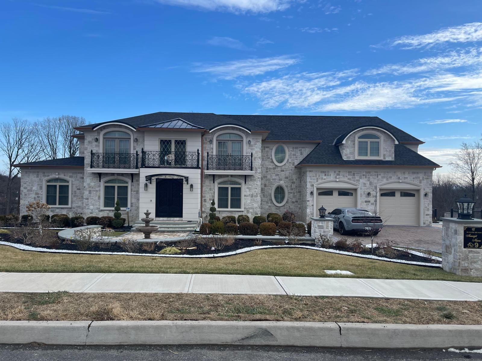 Large two-story house with light stone exterior, black roof, and manicured landscaping under a blue sky.