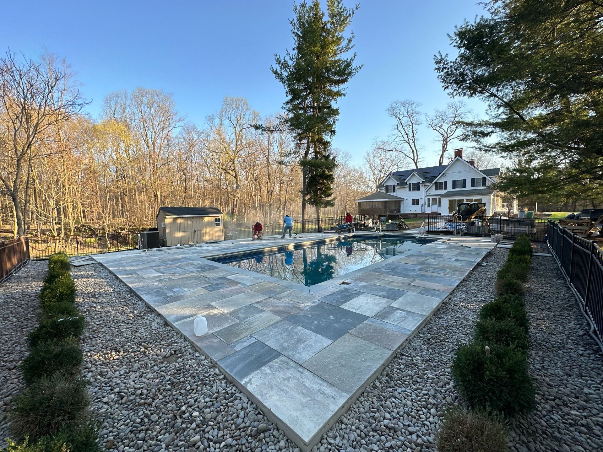 Swimming pool with stone patio, surrounded by trees and a house.