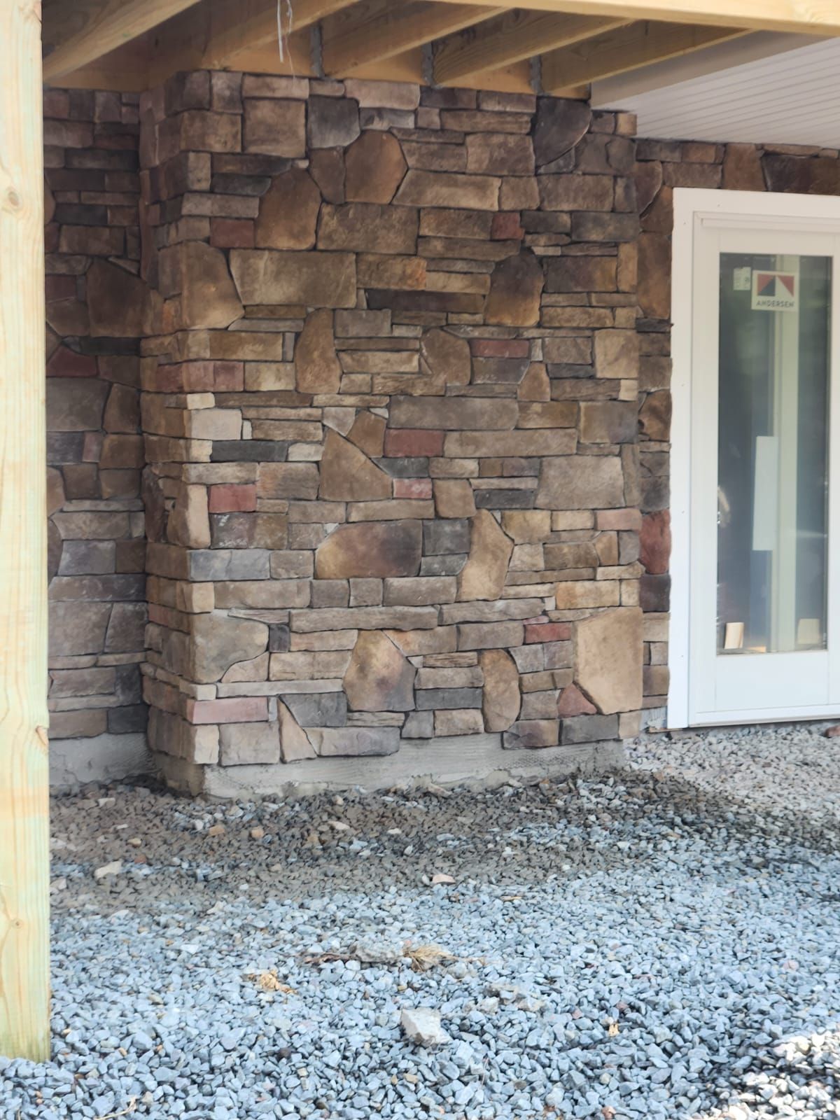 Stone veneer wall under a wooden structure; next to a white door, set on a gravel base.