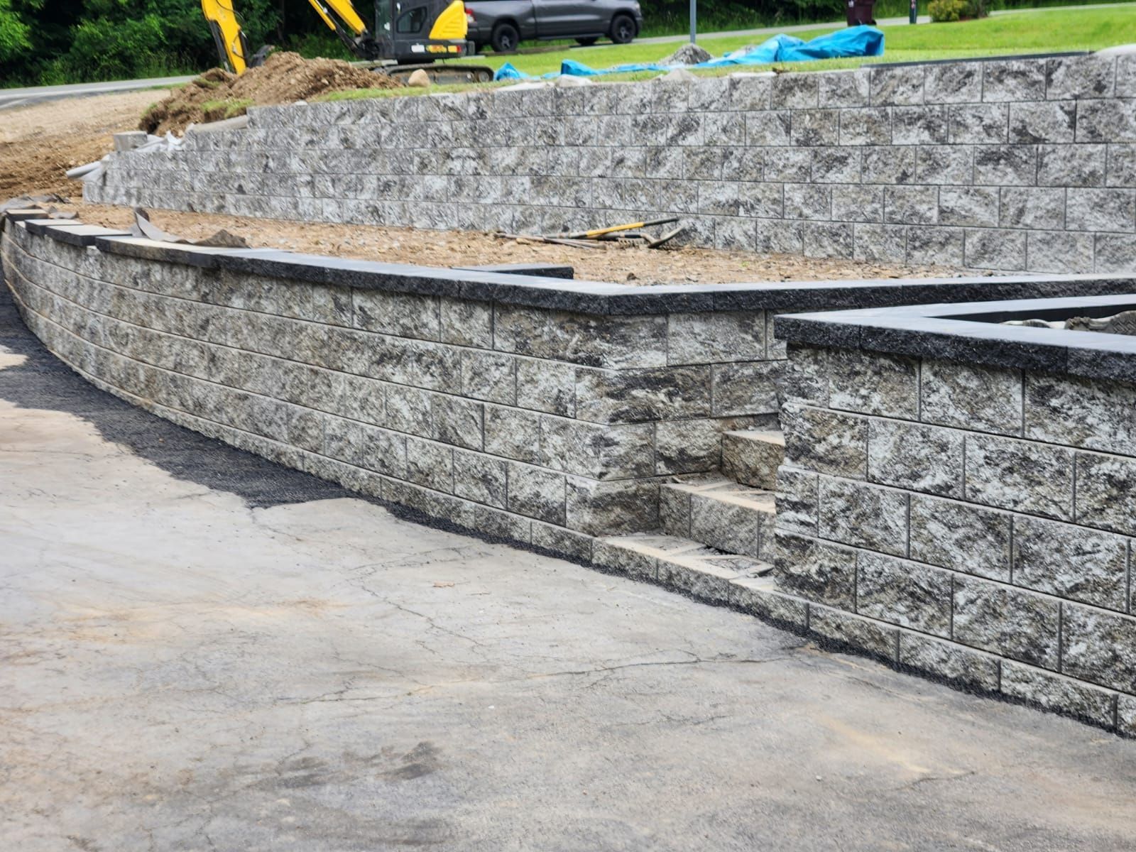 Gray stone retaining wall with steps, curved design, near asphalt, construction equipment in the background.