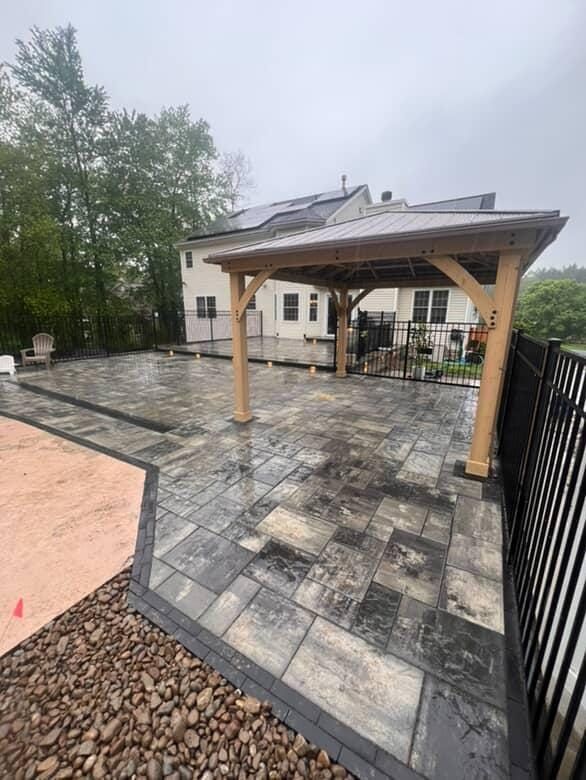 Patio with dark gray pavers, wooden pergola, black fence, and house in background.