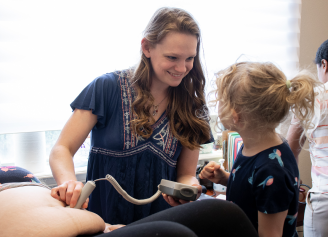 A provider uses a Doppler ultrasound device on a patient's abdomen while a child watches in a brightly lit room.