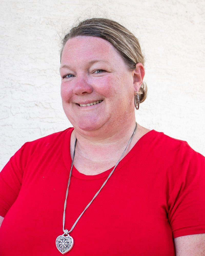 A person wearing a bright red shirt and a heart-shaped pendant necklace smiles against a light, textured background.