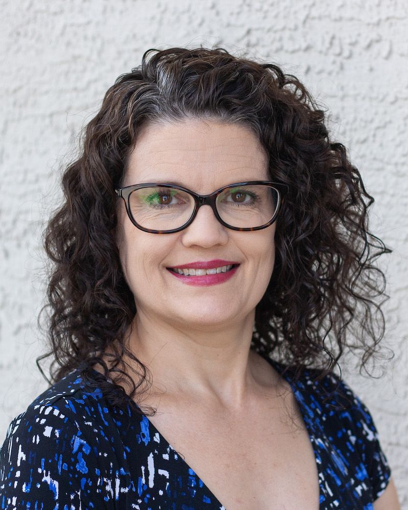 A person with curly dark hair and glasses smiles against a textured white wall, wearing a blue and black patterned top.