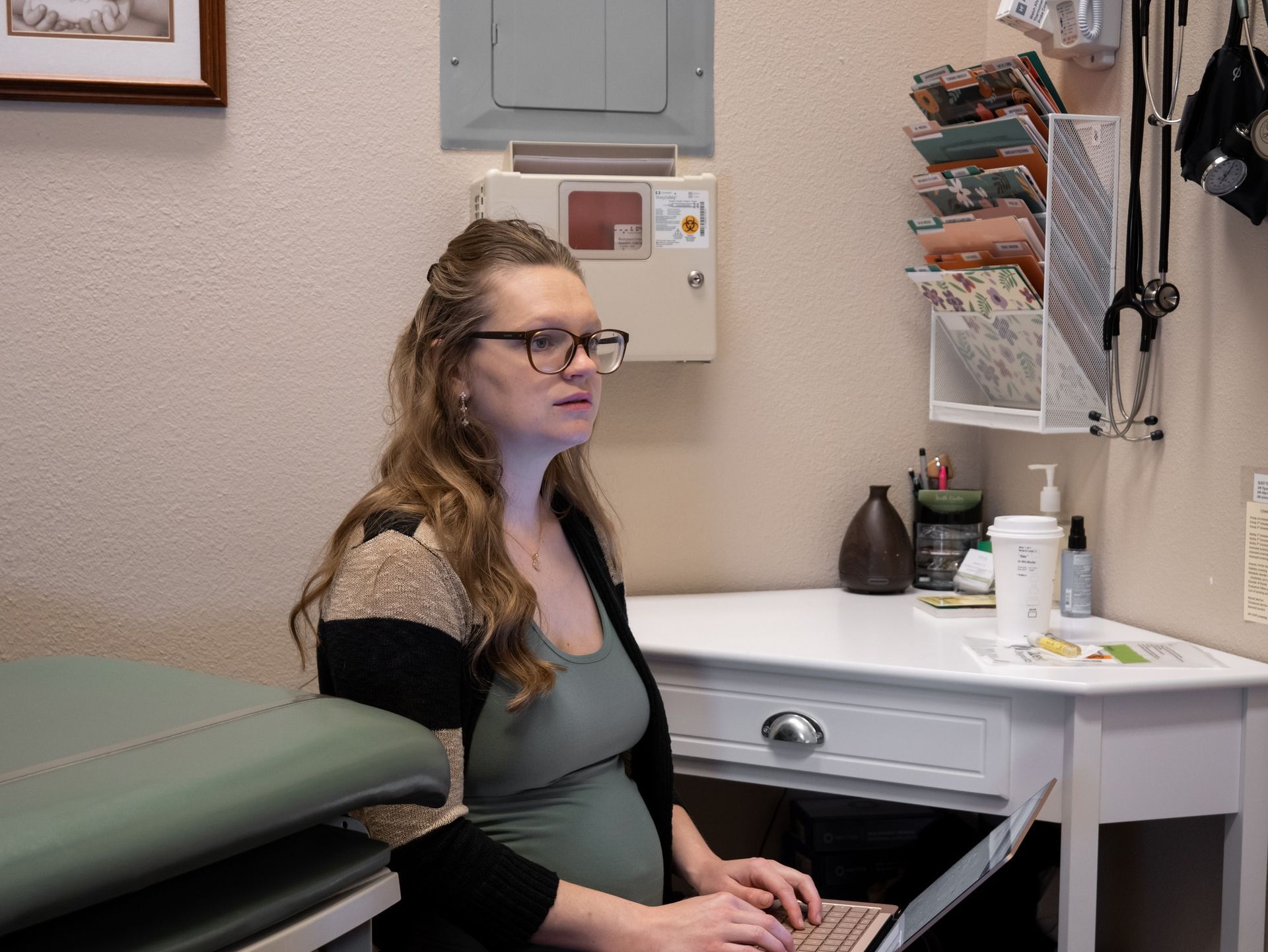 A pregnant person wearing glasses and a cardigan sits at a white desk in a medical office, working on a laptop.