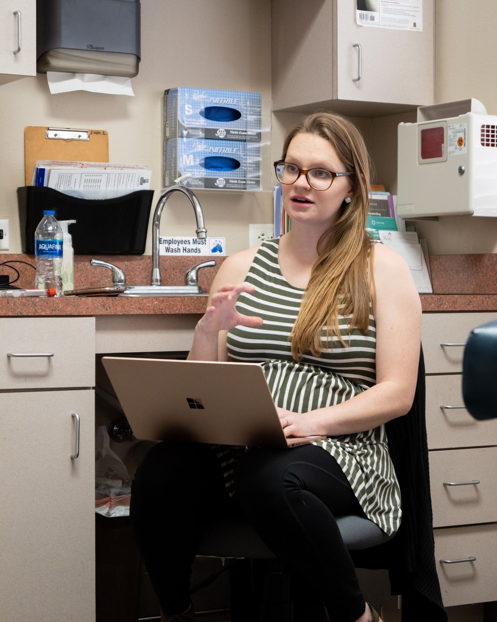 A person wearing glasses and a striped top sits in a medical office, using a laptop and gesturing while speaking.