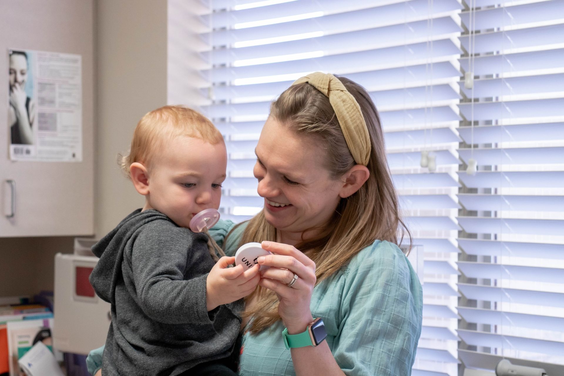 A smiling person holds a toddler who is looking at a small, circular object in a brightly lit office setting.
